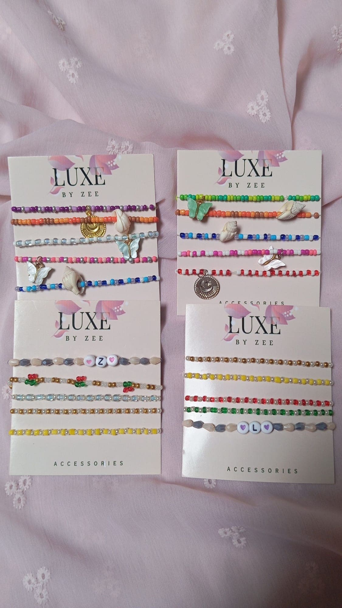 Four Luxe by Zee accessory cards laid on pink fabric, each strung with five beaded bracelets in different palettes
